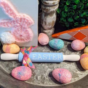 May include: A blue rolling pin with the word "EASTER" written on it, decorated with a pink and white ribbon. The rolling pin is surrounded by glittery pastel-colored eggs on a metal tray.