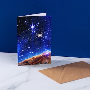 May include: A folded greeting card with a vibrant image of a starry galaxy, featuring bright stars and nebulas in shades of blue, orange, and brown. A brown envelope rests on a white marble surface, with a deep blue background.