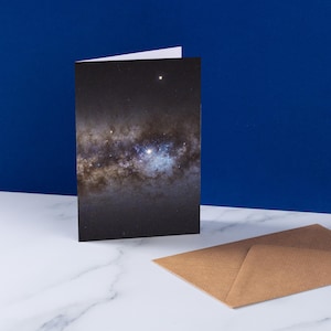 May include: A folded greeting card featuring a detailed image of a galaxy with stars and nebulae. A brown envelope rests on a white marble surface next to the card. The background is a solid blue.