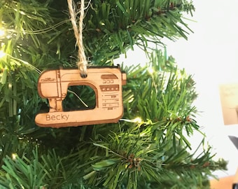 May include: A wooden sewing machine Christmas tree ornament. The light brown ornament has etched details and the name "Becky" engraved on it. The green tree provides a festive backdrop, with a gift box visible in the background.