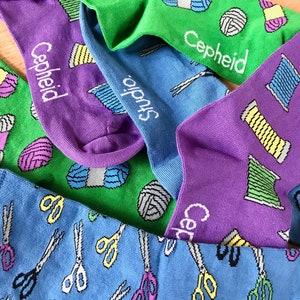 May include: A collection of colorful socks with different patterns. One pair features a green background with yarn balls and the text "Cepheid Studio". Another pair has a purple background with yarn balls and scissors. The third pair has a blue background with scissors.