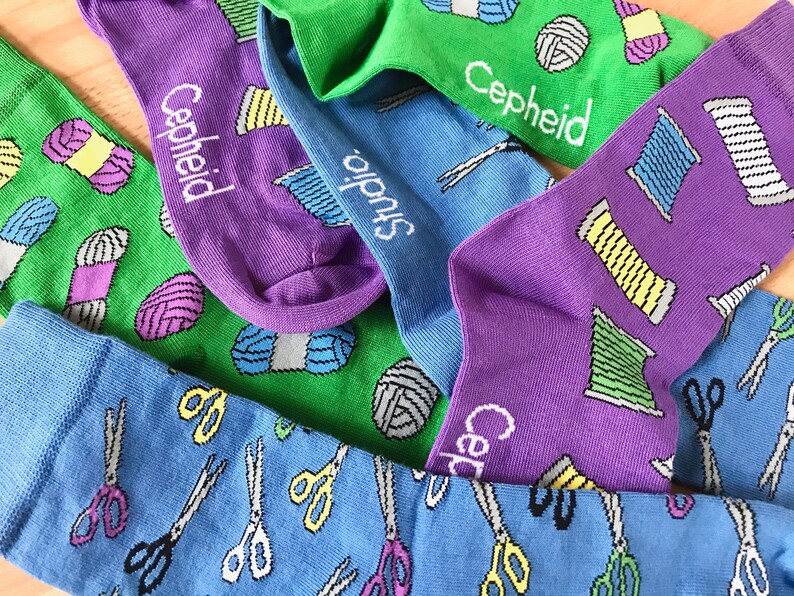 May include: A collection of colorful socks with different patterns. One pair features a green background with yarn balls and the text "Cepheid Studio". Another pair has a purple background with yarn balls and scissors. The third pair has a blue background with scissors.
