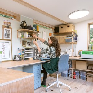 May include: A woman works at a desk in a home office with a laser cutter, shelves, and a large window. The desk is made of wood and has a light blue drawer unit. The woman is wearing a striped shirt and a green skirt.
