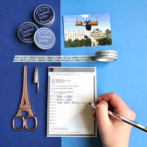 Pu&ograve; includere: Un primo piano di un elenco di cose da fare con compiti scritti a mano, tra cui "Fai un caff&egrave;", "Bevi un caff&egrave;" e "Pensa a iniziare a lavorare". L'elenco &egrave; su un blocco note bianco con un bordo grigio. Ci sono anche tre rotoli di nastro washi con il testo "Cepheid Studio" su di essi, un paio di forbici dorate a forma di Torre Eiffel, una penna d'argento e un cappuccio di penna d'argento.