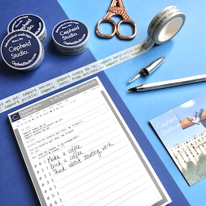 May include: A white notebook with gray lines and a gray border. The notebook has the text "Cepheid Studio Notebooks" printed on the cover. The notebook is open to a page with handwritten notes and code. The code includes the text "import numpy as np: import matplotlib.pyplot as plt: import scipy.stats as st: import astropy.io.fits as fits: import jax.numpy as jnp: import tensorflow as tf". The notebook is on a blue surface with a roll of washi tape, a pen, and a pair of scissors.