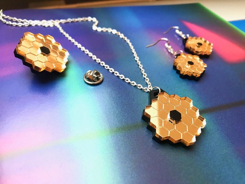 May include: A gold necklace with a hexagonal pendant, a matching pair of earrings, and a gold pin. The pendant and earrings are made of mirrored acrylic and have a black center.