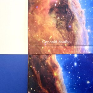 May include: A section of a fabric with a celestial nebula design in shades of orange, brown, and blue. The text "Cepheid Studio" is printed in white. The fabric is bordered by white and blue sections.