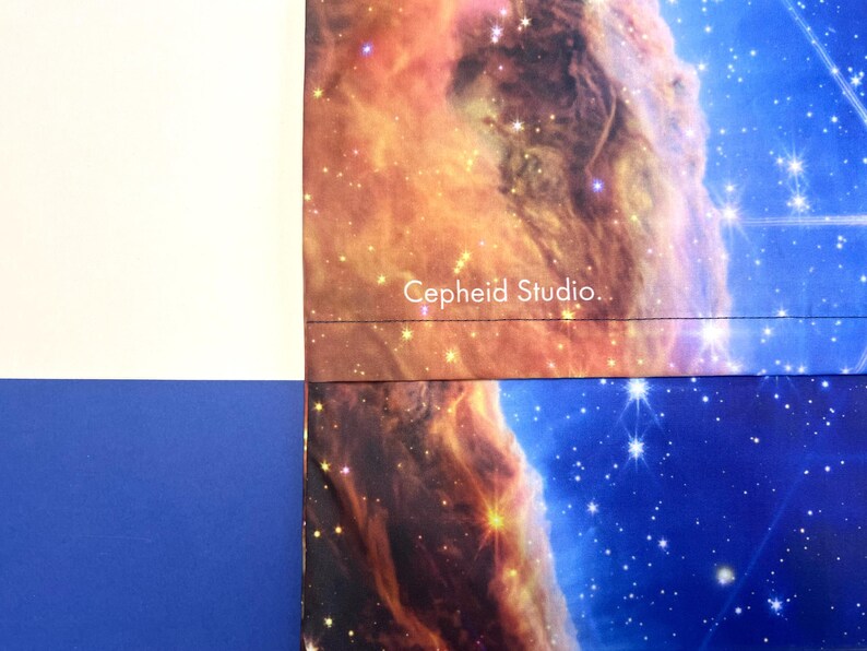 May include: A section of a fabric with a celestial nebula design in shades of orange, brown, and blue. The text "Cepheid Studio" is printed in white. The fabric is bordered by white and blue sections.