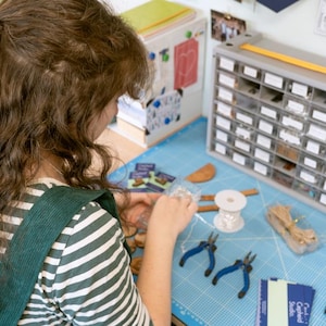 May include: A person is working on a craft project at a table. They are using a variety of tools and materials, including pliers, wire, beads, and a spool of thread. There are also several small boxes filled with various items. The table is covered in a blue mat with a grid pattern.