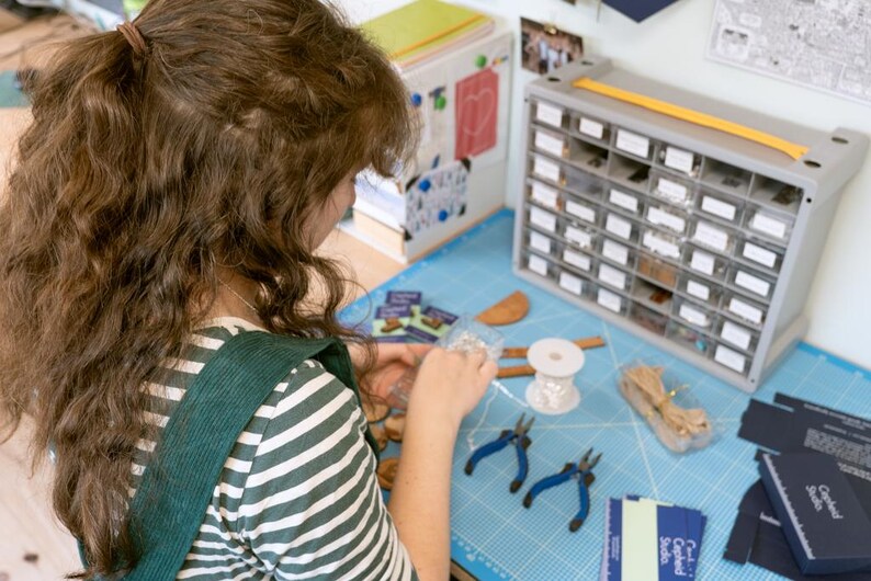 May include: A person is working on a craft project at a table. They are using a variety of tools and materials, including pliers, wire, beads, and a spool of thread. There are also several small boxes filled with various items. The table is covered in a blue mat with a grid pattern.
