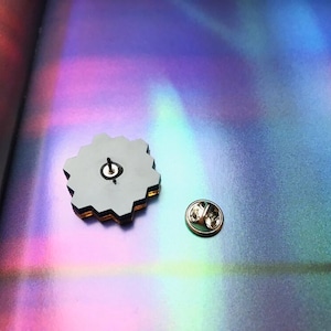 May include: A hexagonal-shaped pin with a black and gold design. The pin is on a holographic surface with a rainbow effect. The pin has a silver back with a pin post.