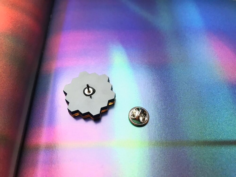 May include: A hexagonal-shaped pin with a black and gold design. The pin is on a holographic surface with a rainbow effect. The pin has a silver back with a pin post.