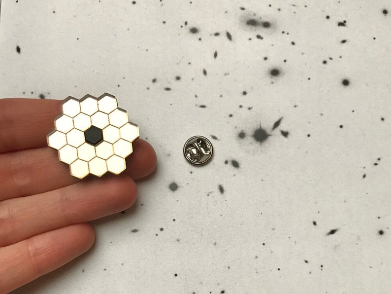 May include: A gold-colored pin with a black center and a honeycomb design. The pin is on a white background with black speckles.