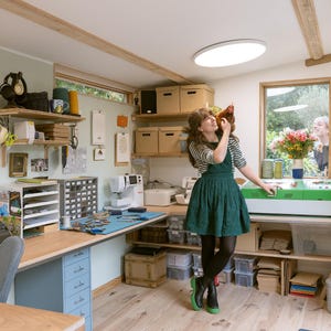 May include: A well-lit craft room with a woman holding a chicken. The room features a sewing machine, storage boxes, and a large wooden desk. Shelves display various craft supplies and tools. The woman is wearing a green dress and black boots.