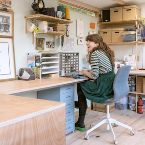 May include: A woman sits at a desk in a workshop, working on a project. The desk is made of wood and has a light blue drawer unit. The woman is wearing a green dress and a striped shirt. There are shelves on the wall behind her, filled with various items.