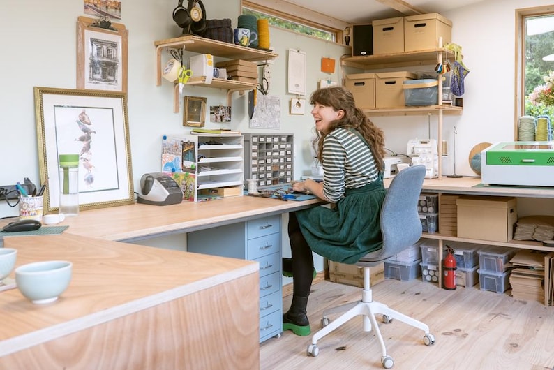 May include: A woman sits at a desk in a workshop, working on a project. The desk is made of wood and has a light blue drawer unit. The woman is wearing a green dress and a striped shirt. There are shelves on the wall behind her, filled with various items.