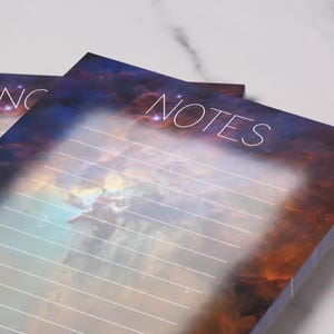 May include: Two notepads with a celestial design. The pads feature a vibrant nebula background in shades of orange, blue, and purple. The word "NOTES" is written in white script at the top. Horizontal lines are visible for writing.