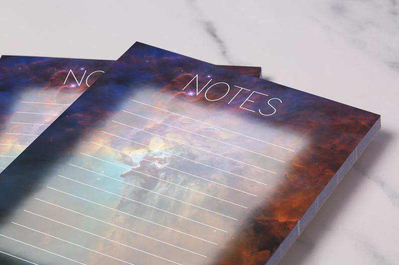 May include: Two notepads with a celestial design. The pads feature a vibrant nebula background in shades of orange, blue, and purple. The word "NOTES" is written in white script at the top. Horizontal lines are visible for writing.