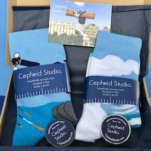 Könnte beinhalten: Zwei Paar Socken in einer Geschenkbox. Ein Paar ist blau mit einem weißen Wolkenmuster und das andere Paar ist weiß mit einem blauen Wolkenmuster. Beide Paare haben den Text "Cepheid Studio. Scientifically accurate socks Designed in Paris Made in the EU" auf der Verpackung. Die Socken werden von zwei kleinen runden Dosen mit astronomischem Waschband begleitet, jede mit dem Text "Cepheid Studio. CepheidStudio.com" auf dem Etikett.