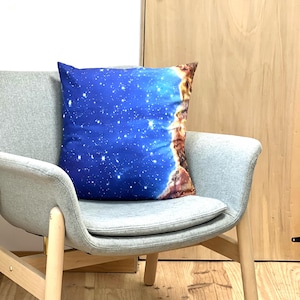 May include: A square decorative pillow with a celestial design. The pillow features a blue background with white stars and a brown and orange nebula-like design on the right side. It rests on a gray chair with wooden legs.