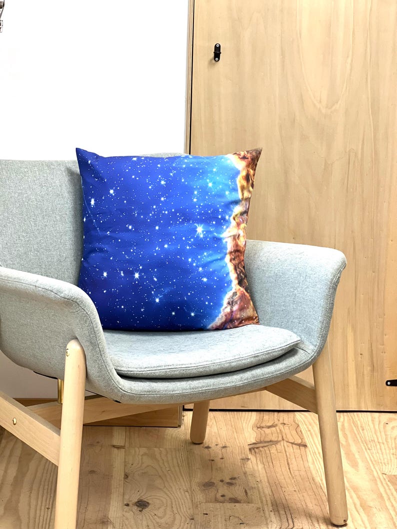 May include: A square decorative pillow with a celestial design. The pillow features a blue background with white stars and a brown and orange nebula-like design on the right side. It rests on a gray chair with wooden legs.