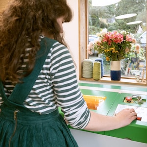 May include: A person operating a green and white machine with a red button. The machine has a digital display and a clear plastic container. The person is wearing a green and white striped shirt and a green overall dress.