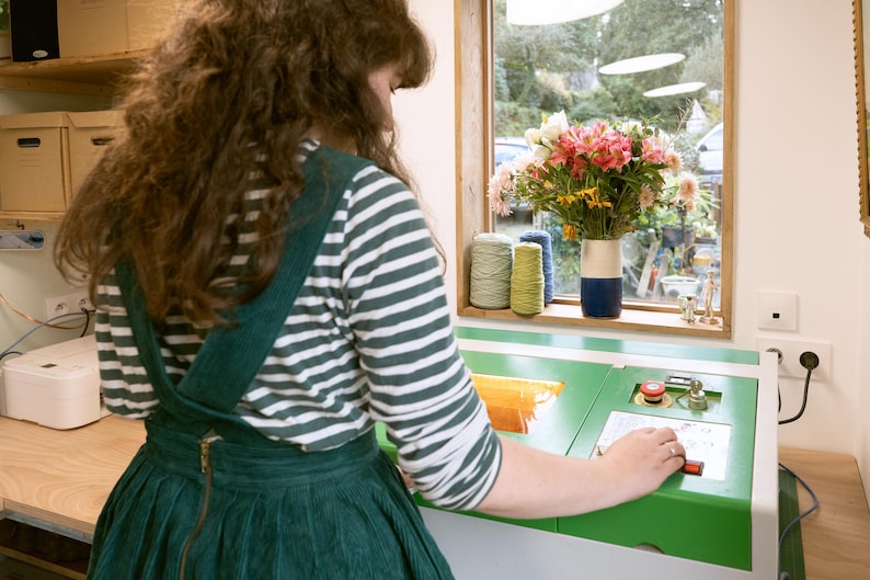 May include: A person operating a green and white machine with a red button. The machine has a digital display and a clear plastic container. The person is wearing a green and white striped shirt and a green overall dress.