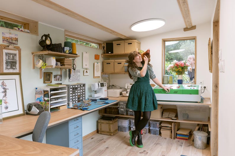 May include: A woman in a green dress holds a brown chicken in a workshop with a sewing machine, shelves, and a laser cutter.