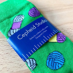 May include: Green socks with a colorful yarn ball pattern. The socks are folded and have a blue label that reads "Cepheid Studio. Perfectly crafted crafty socks. Designed in Paris."