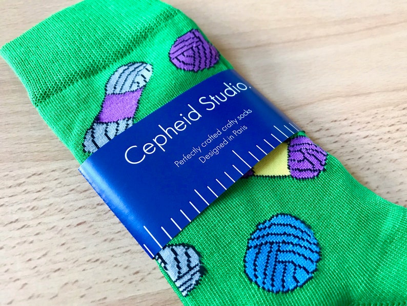 May include: Green socks with a colorful yarn ball pattern. The socks are folded and have a blue label that reads "Cepheid Studio. Perfectly crafted crafty socks. Designed in Paris."