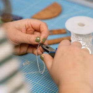 May include: A person uses pliers to attach a silver chain to a piece of jewelry. The chain is on a blue grid surface.