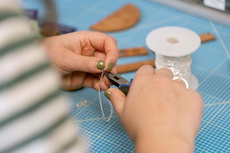 May include: A person uses pliers to attach a silver chain to a piece of jewelry. The chain is on a blue grid surface.