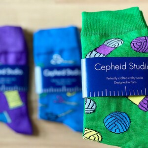 May include: Three pairs of colorful socks with a yarn and needle design. The green socks have a tag that reads "Cepheid Studio. Perfectly crafted crafty socks Designed in Paris."