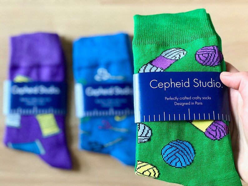 May include: Three pairs of colorful socks with a yarn and needle design. The green socks have a tag that reads "Cepheid Studio. Perfectly crafted crafty socks Designed in Paris."