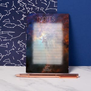 Op de afbeelding: Een notitieblok met een nevelontwerp en het woord "NOTES" bovenaan. Het notitieblok heeft gelinieerde pagina's en een donkere achtergrond. Een roségouden pen ligt op een wit marmeren oppervlak voor het notitieblok.