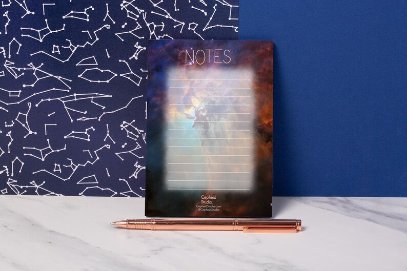 May include: A notepad with a nebula design and the word "NOTES" at the top. The notepad has lined pages and a dark background. A rose gold pen rests on a white marble surface in front of the notepad.