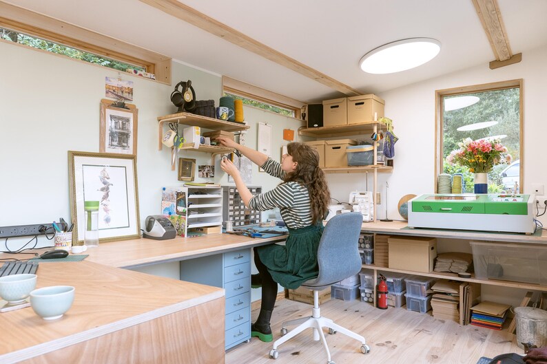 May include: A woman works at a desk in a home office with a laser cutter, shelves, and a large window. The desk is made of wood and has a light blue drawer unit. The woman is wearing a striped shirt and a green skirt.