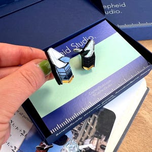 Op de afbeelding: Een paar manchetknopen met een geometrisch, veelzijdig ontwerp met zwarte, witte en goudkleurige accenten. De manchetknopen worden gepresenteerd in een donkerblauwe doos met het label Cepheid Studio, inclusief een liniaal langs de rand.