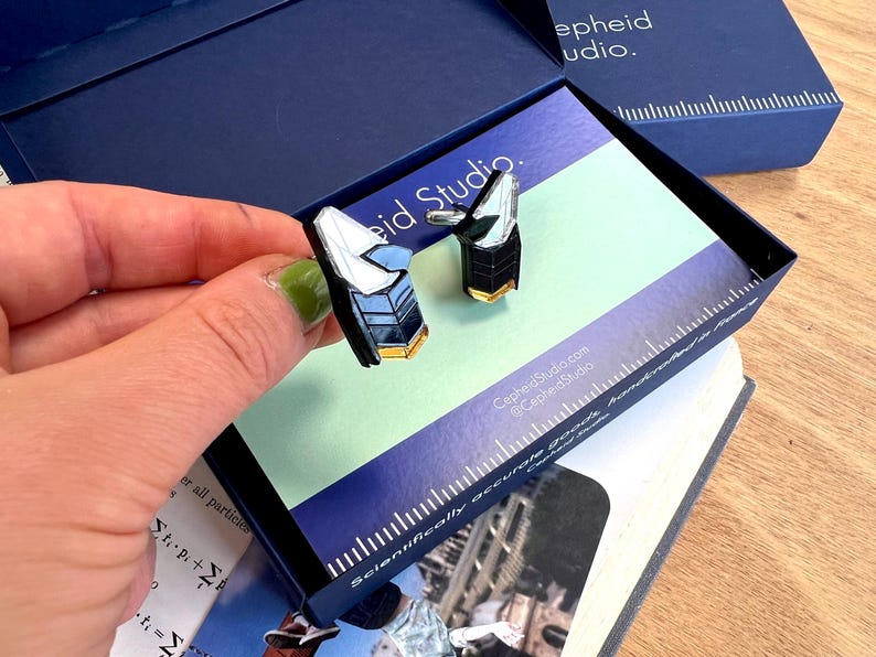Op de afbeelding: Een paar manchetknopen met een geometrisch, veelzijdig ontwerp met zwarte, witte en goudkleurige accenten. De manchetknopen worden gepresenteerd in een donkerblauwe doos met het label Cepheid Studio, inclusief een liniaal langs de rand.