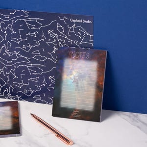 May include: Stationery set featuring a celestial theme. Includes a large navy blue print with white constellation outlines, a notepad with the word "NOTES", and a small notepad. A rose gold pen rests on the surface. The text "Cepheid Studio" is visible.