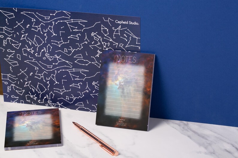 May include: Stationery set featuring a celestial theme. Includes a large navy blue print with white constellation outlines, a notepad with the word "NOTES", and a small notepad. A rose gold pen rests on the surface. The text "Cepheid Studio" is visible.