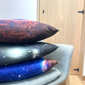 May include: Three decorative pillows with celestial designs are stacked on a gray chair. The top pillow has a reddish-brown nebula pattern, the middle one a black and white galaxy, and the bottom one a blue starry sky.