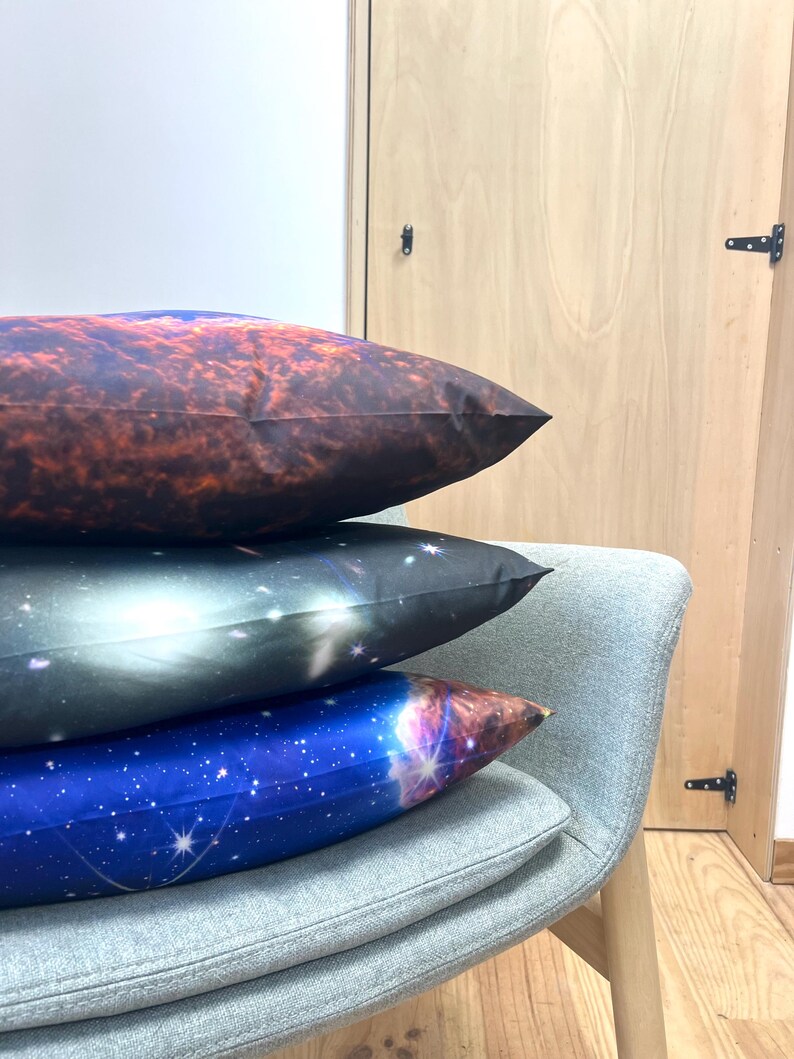 May include: Three decorative pillows with celestial designs are stacked on a gray chair. The top pillow has a reddish-brown nebula pattern, the middle one a black and white galaxy, and the bottom one a blue starry sky.
