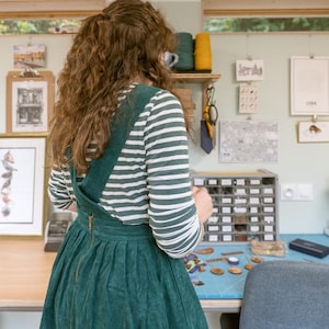 May include: A woman wearing a green and white striped shirt and a green corduroy overall dress is standing in a craft room. She is looking at a small box of colorful buttons. The room has a sewing machine, a table with a variety of craft supplies, and several framed pictures on the walls.