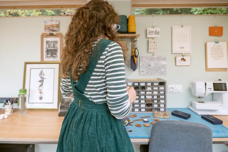 May include: A woman wearing a green and white striped shirt and a green corduroy overall dress is standing in a craft room. She is looking at a small box of colorful buttons. The room has a sewing machine, a table with a variety of craft supplies, and several framed pictures on the walls.