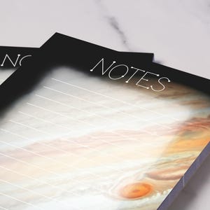 Puede incluir: Dos blocs de notas negros con la palabra "NOTES" en escritura blanca. El fondo de los blocs de notas presenta una imagen colorida de Júpiter. Los blocs de notas tienen páginas rayadas blancas.