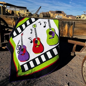 Może przedstawiać: Kolorowa kołdra z motywem muzycznym przedstawiająca trzy gitary w kolorze fioletowym, czerwonym i zielonym z czarnymi i białymi nutami. Kołdra leży na drewnianym kole wozu.