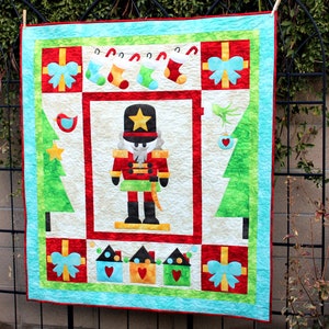 Könnte beinhalten: Eine farbenfrohe Weihnachtsdecke mit einem Nussknacker, Weihnachtsbäumen, Geschenken und Strümpfen. Die Decke ist aus verschiedenen Stoffen gefertigt und hat ein Patchwork-Design.