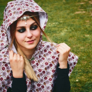 May include: A woman wearing a white and burgundy hooded cloak with a floral pattern. She is wearing a silver headband with a pearl pendant. The cloak has a white lining and is made of a soft, flowing fabric.