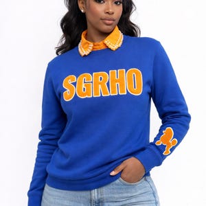May include: A royal blue sweatshirt with the letters "SGRHO" in orange across the chest. The sweatshirt has an orange collar with white pearl-like embellishments and an orange poodle graphic on the left sleeve.